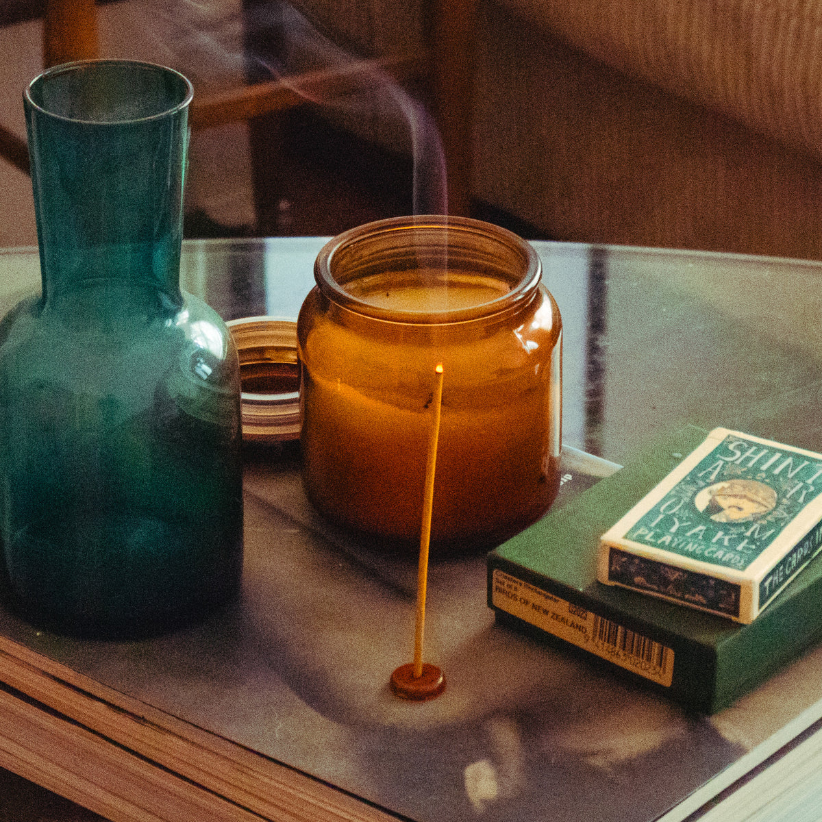 A warm, vintage display of a teal glass vase, amber candle jar, incense stick, and green matchbox on a wooden table, evoking calm nostalgia.