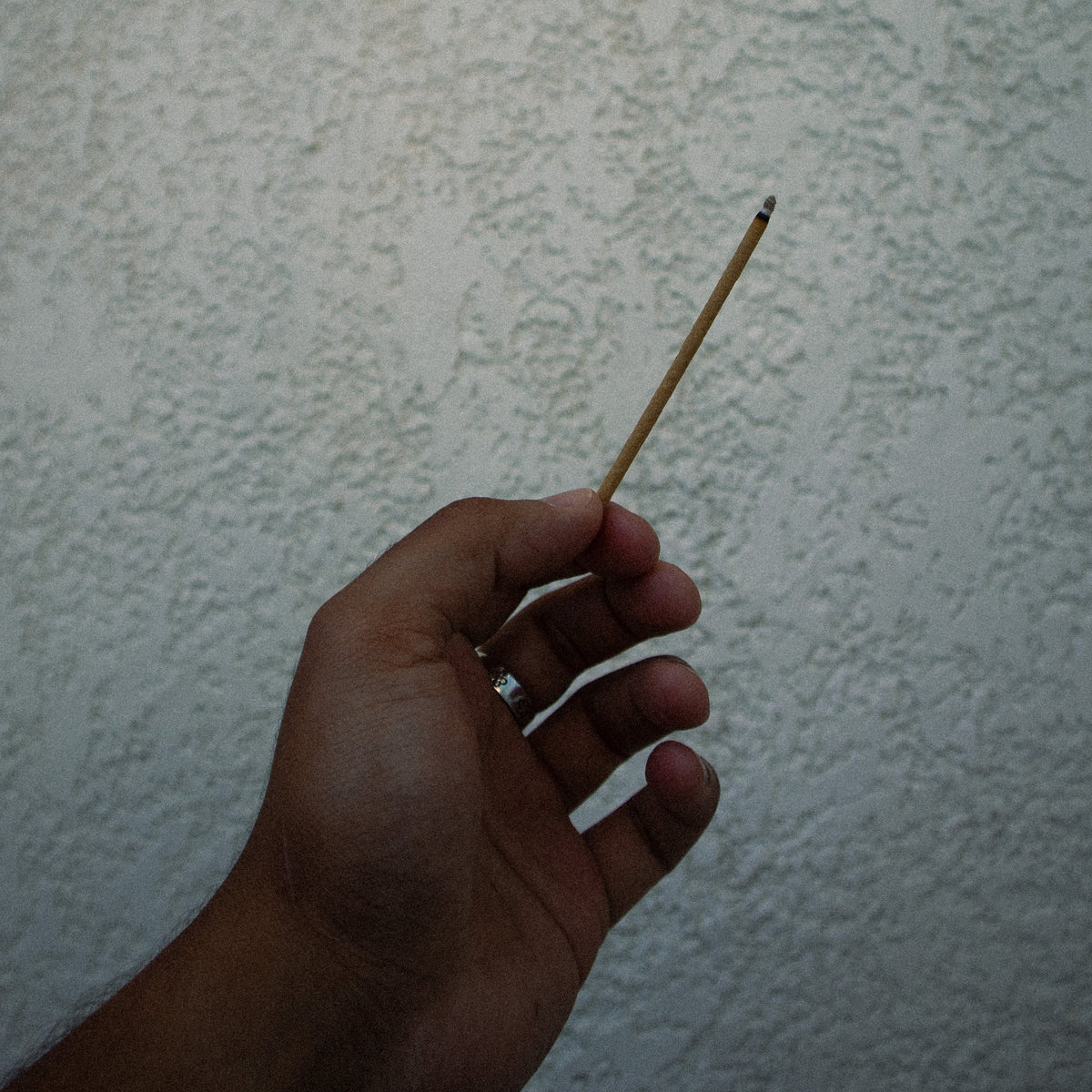 A hand holding a thin incense stick against a textured off-white wall.
