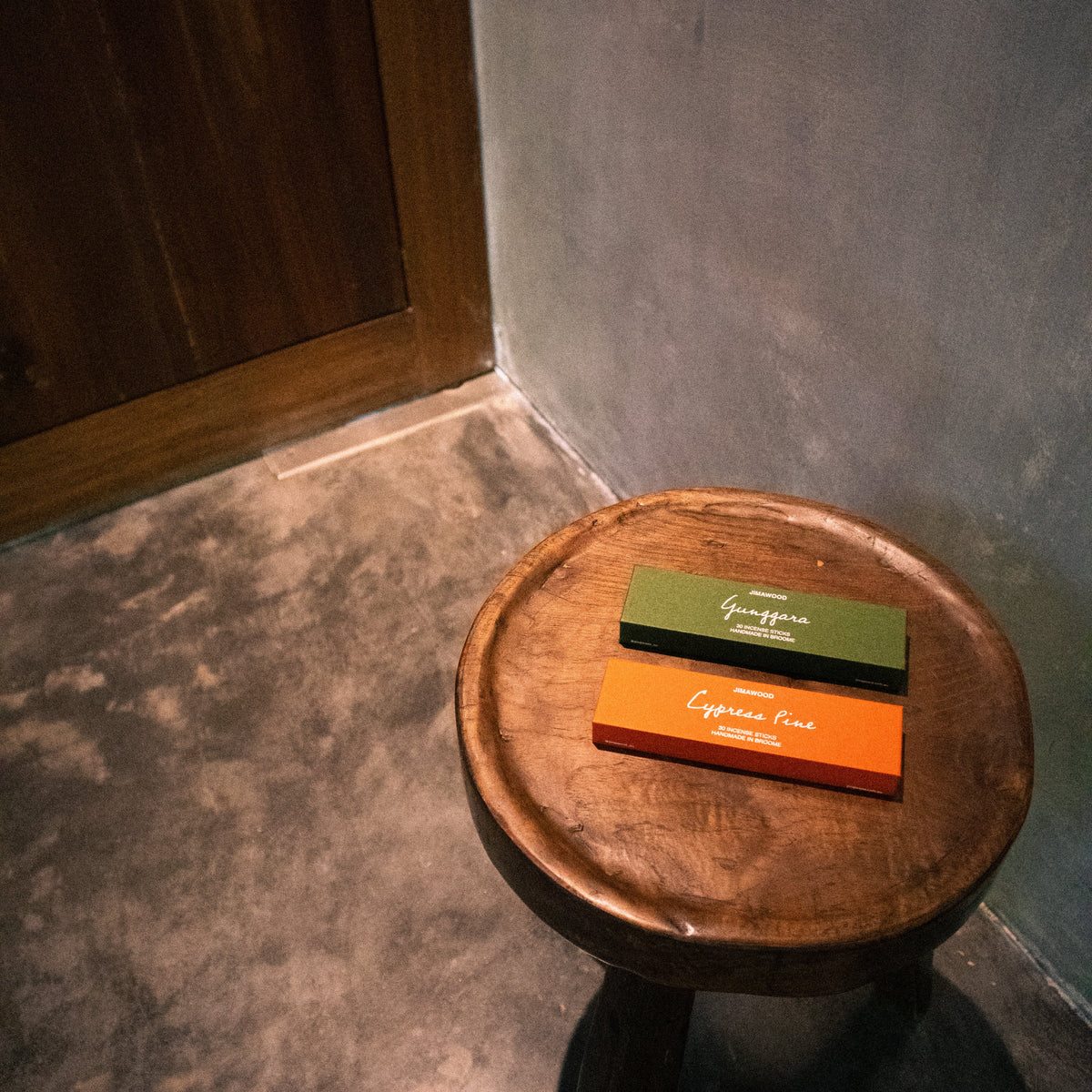Two rectangular boxes, one green and one orange, stacked on a round wooden stool in a dimly lit room with a textured gray wall and concrete floor.
