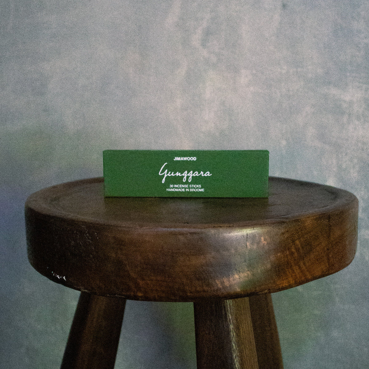 a green box atop a wooden stool against grey wall. the box says: jimawood. Gunggara. 30 incense sticks, handmade in broome