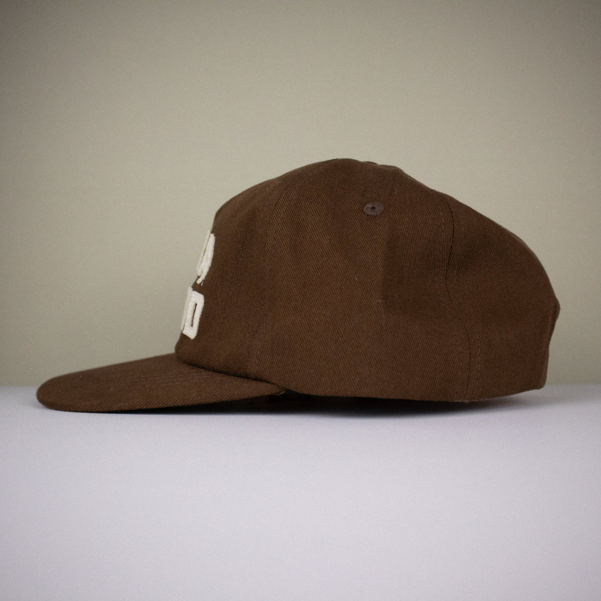 A brown flat-brim baseball cap with a white embroidered logo on the side, displayed on a white surface against a neutral gray background.