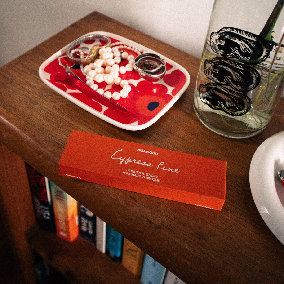 An orange box of Jimawood incense which is an orange colour sitting on a bookshelf slightly askew. There are other objects on the shelf also including jewellery on a red dish and a botlle with water in it.