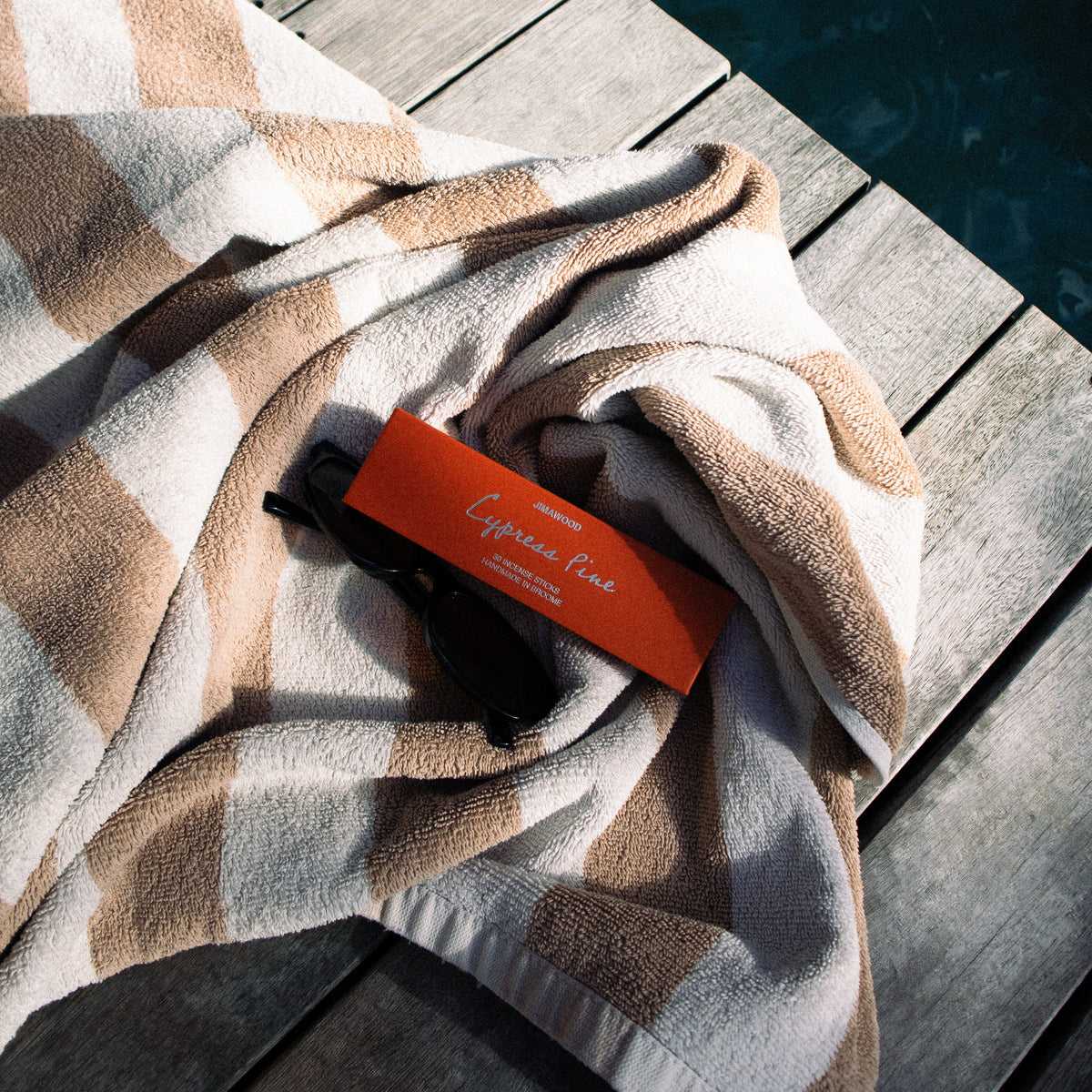 A beige and white checkered towel draped on a weathered wooden deck next to dark blue water, with a small orange tag reading 'Cypress Pine' resting on it.