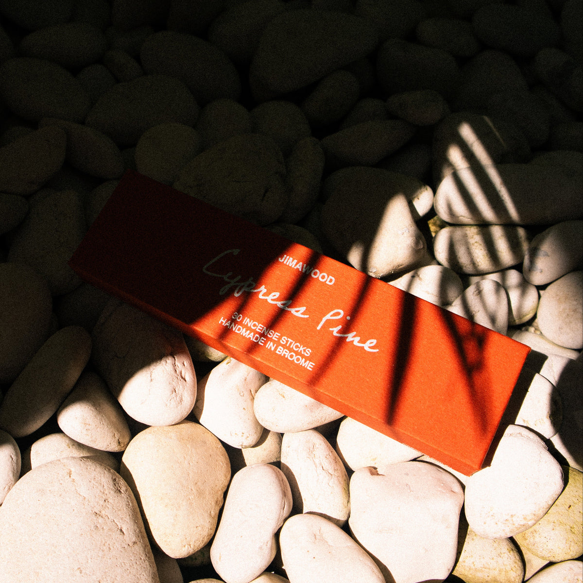 A rectangular orange-red product box with white script text lies on smooth white stones outdoors in strong sunlight and deep shadow.