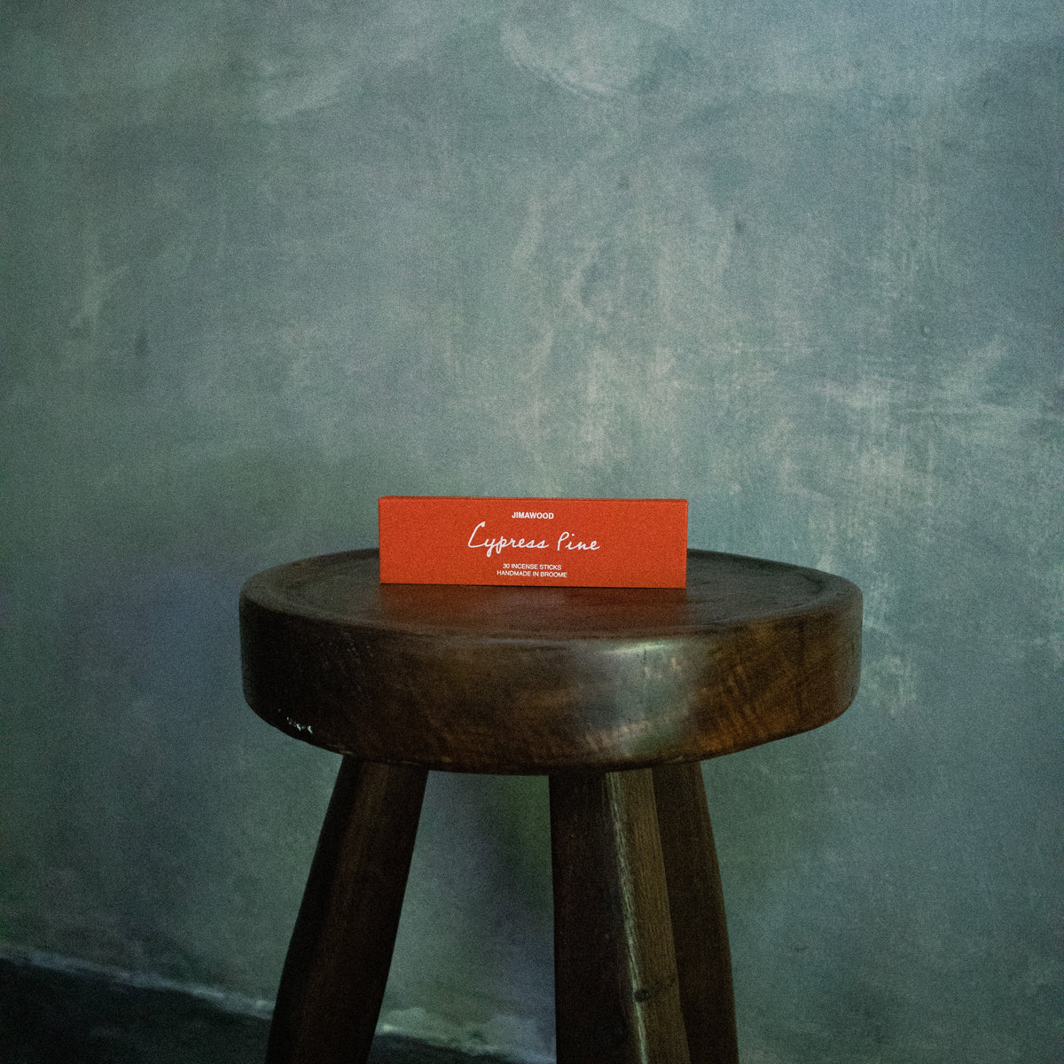 A small orange rectangular box with white script text sits on the top of a dark wooden stool against a textured gray wall.