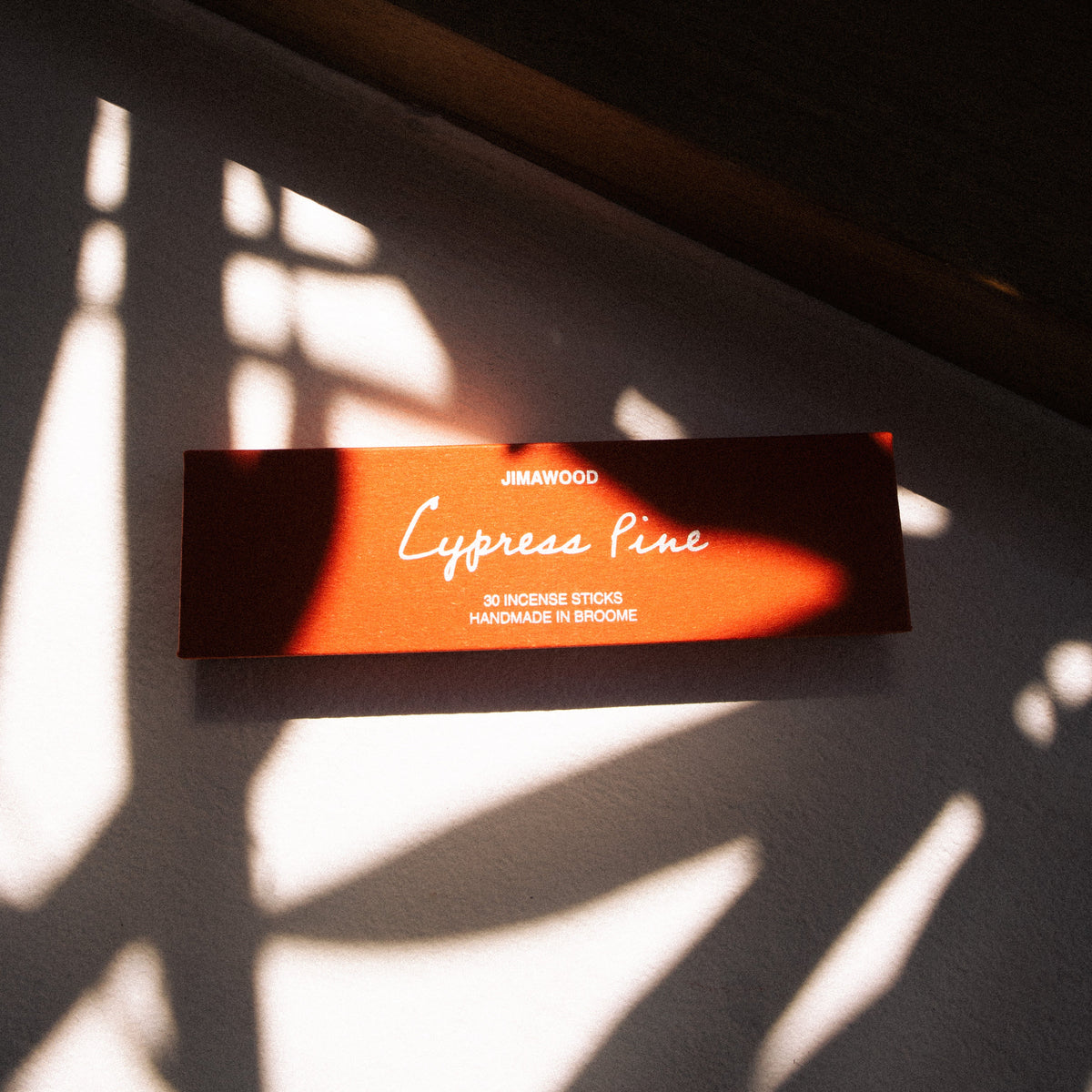 A shadowy image of an orange box near w window sill. the box says: jimawood. Cypress Pine. 30 Incense Sticks. Handmade in Broome
