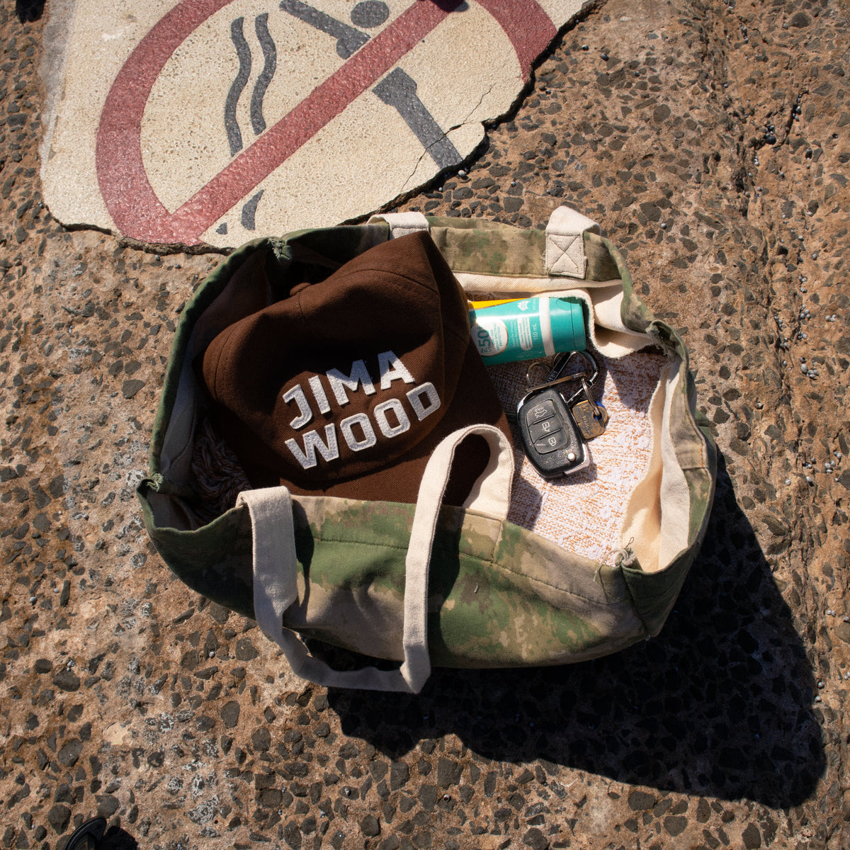 A camouflage canvas tote bag sits on a textured concrete surface, filled with a brown baseball cap featuring the text 'JIMA OOD' in white, a tube of sun screen and a black car key fob.