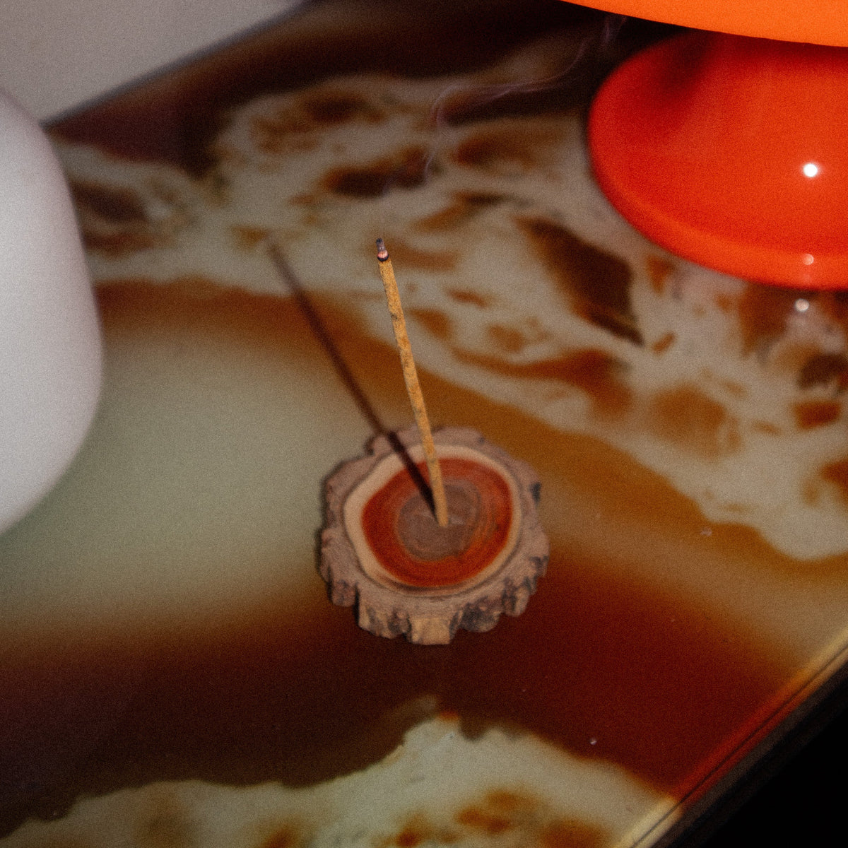 A close-up of a small incense holder with a single burning incense stick, placed on a glossy brown and cream marbled surface beneath a large orange lamp.